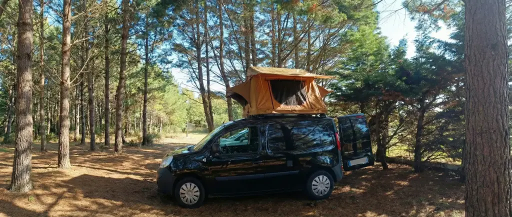 Un Renault Kangoo noir avec une tente de toit Swisskings couleur orange et marron installée et ouverte, stationné dans une forêt de pins ensoleillée avec les portes arrière ouvertes.